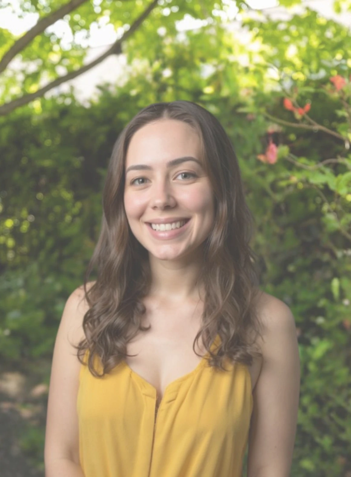 A woman in a yellow top smiles warmly, standing in a lush garden with green foliage and red flowers, conveying a cheerful and serene mood.